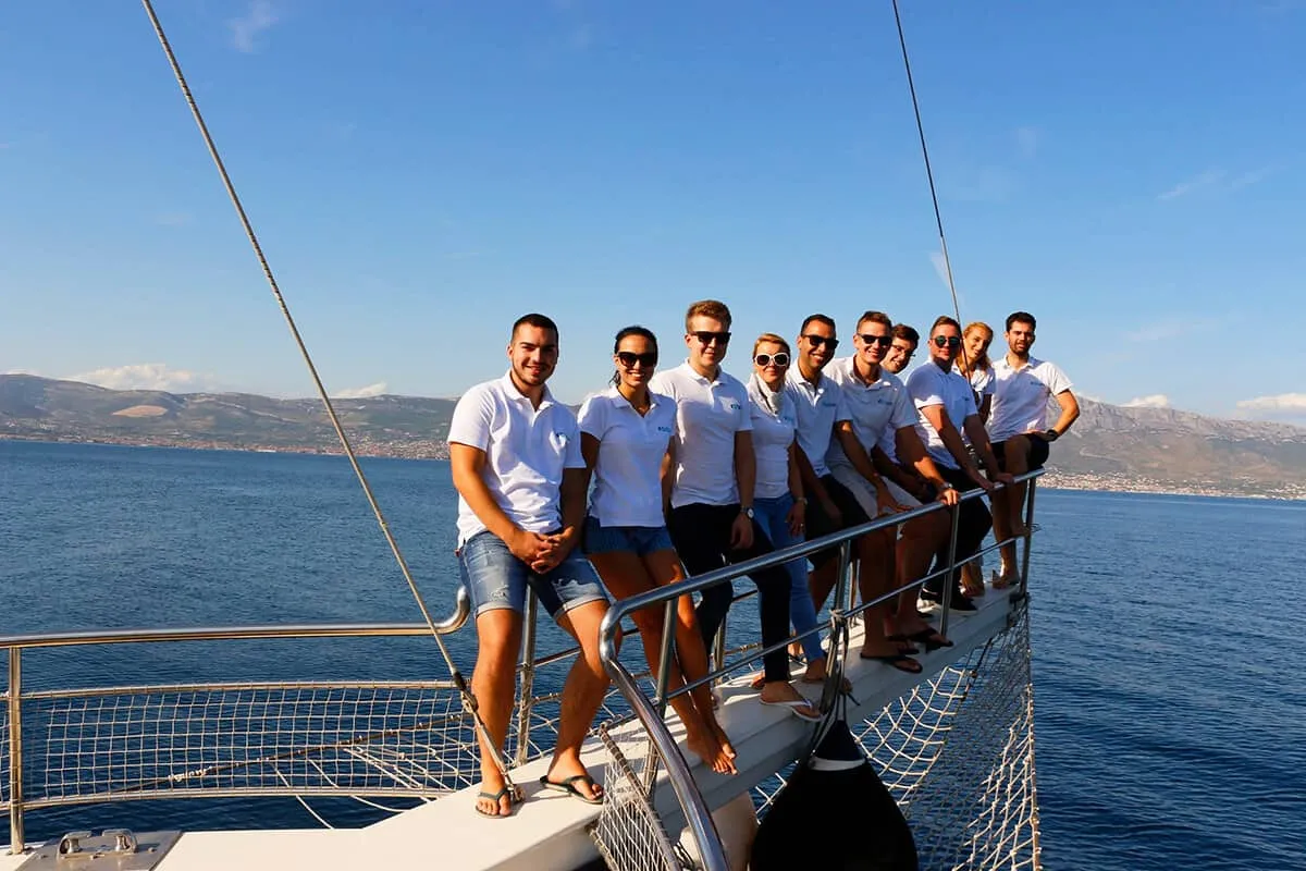 A group of people in white shirts and sunglasses stand together on the bow of a sailboat, smiling, with calm blue sea and distant coastline in the background.