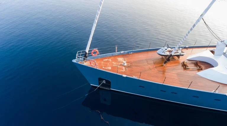 Aerial view of the bow of a modern yacht with a wooden deck floating on calm, deep blue water. A jet ski is secured on the deck, and a red lifebuoy hangs on the railing.