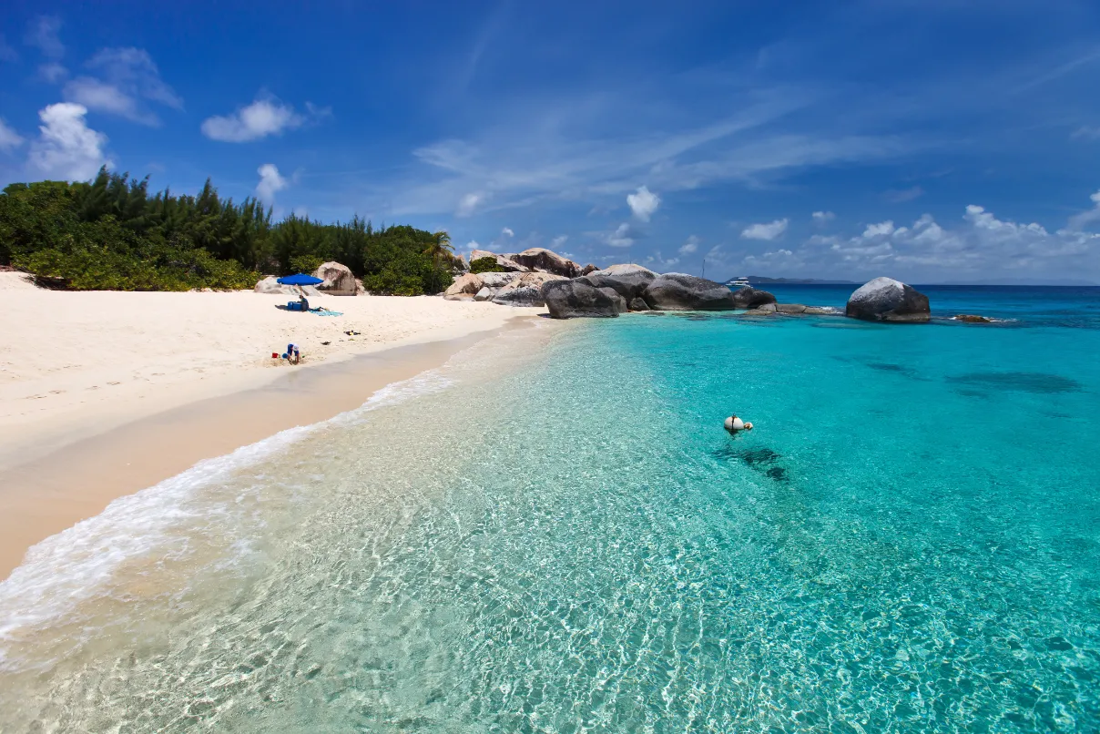 A tropical beach with clear turquoise water, white sand, scattered rocks, and lush green bushes under a bright blue sky with wispy clouds. Two people relax on the sand near the water’s edge.