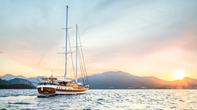 A large sailboat with two masts sails on a calm sea at sunset, with mountains in the background and the sun low on the horizon, casting a warm glow over the water.