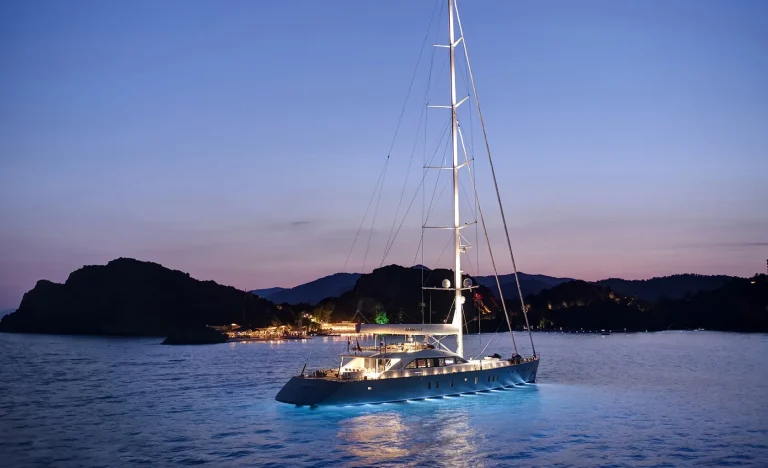 A sleek, modern yacht with glowing underwater lights floats on calm water at dusk, with silhouetted mountains and a softly lit shoreline in the background.