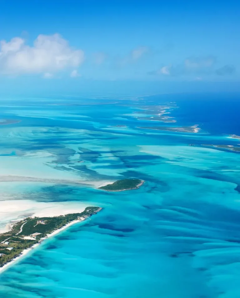 Aerial view of a tropical coastline with small islands, clear turquoise water, white sandy beaches, and patches of greenery under a blue sky with scattered clouds.