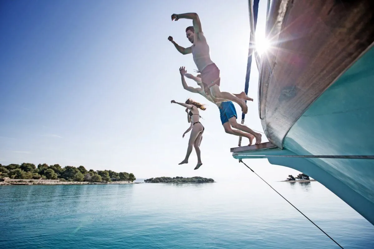 Four people jump off the side of a boat into clear blue water on a sunny day, with a small island and trees visible in the background. The scene captures a moment of adventure and fun.