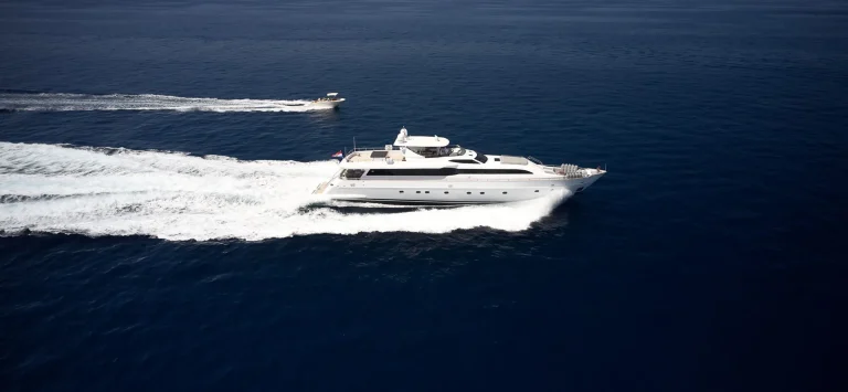 A large white luxury yacht cruises quickly on deep blue water, leaving a trail of white foam behind. A smaller motorboat follows close by, both moving across the calm sea under a clear sky.