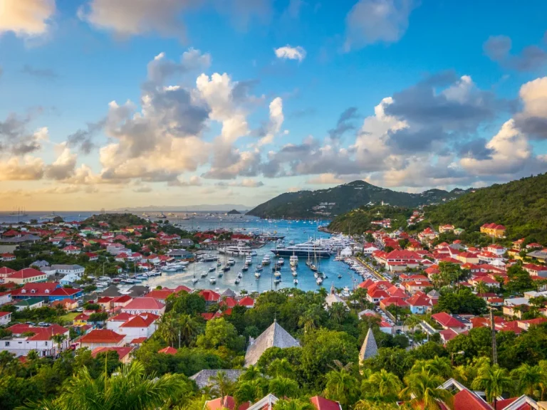 A scenic view of a coastal town with red-roofed buildings, lush green hills, and a harbor filled with boats under a partly cloudy sky at sunset.
