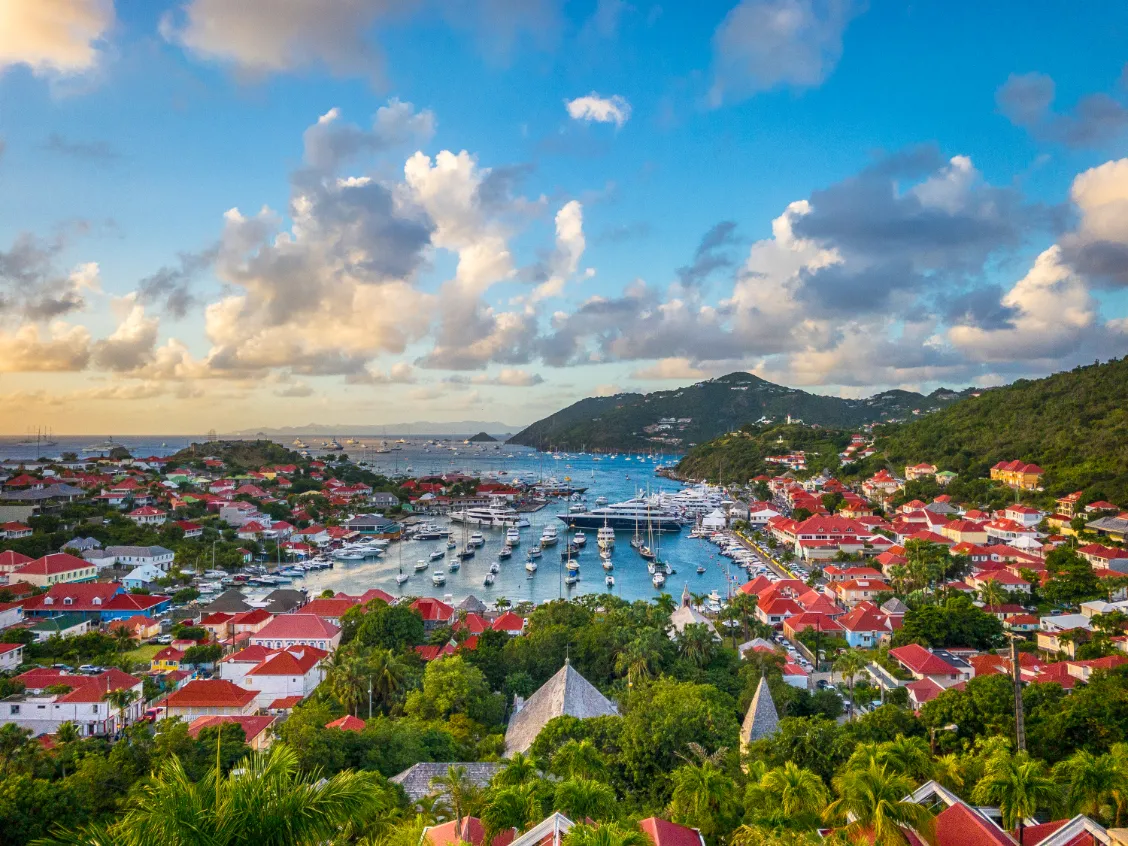 A scenic view of a coastal town with red-roofed buildings, lush green hills, and a harbor filled with boats under a partly cloudy sky at sunset.