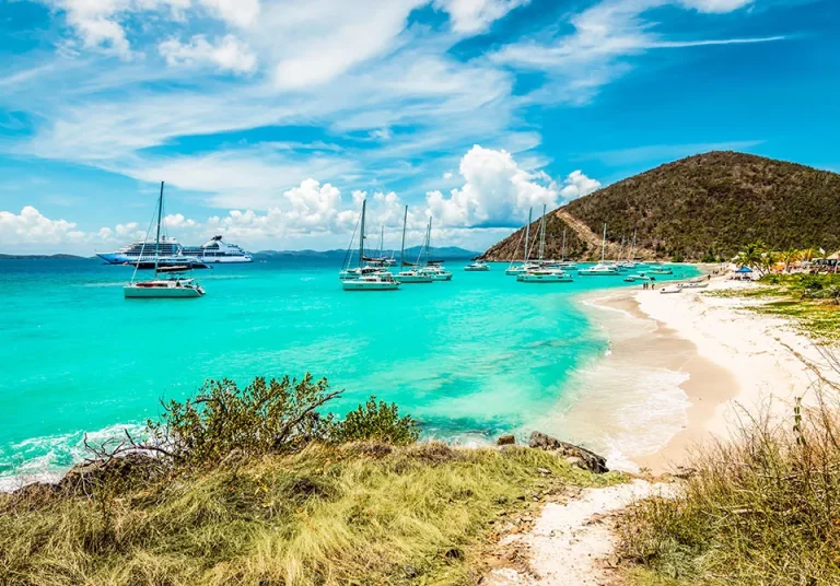 Turquoise water with yachts and sailboats anchored near a sandy beach. A grassy path leads to the shore, with lush hills and palm trees in the background under a bright, partly cloudy sky.