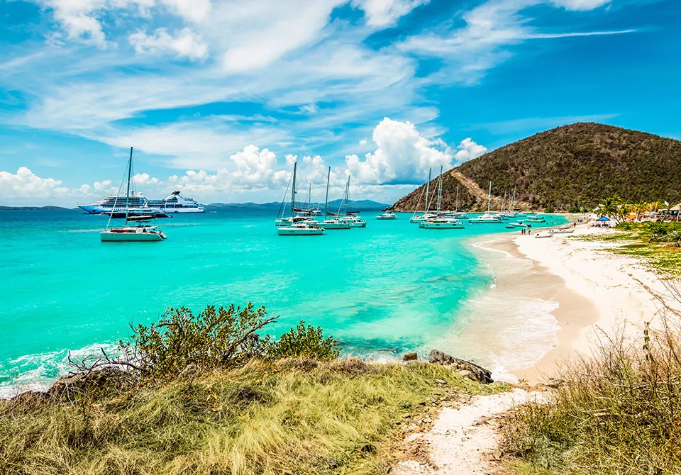 Turquoise water with yachts and sailboats anchored near a sandy beach. A grassy path leads to the shore, with lush hills and palm trees in the background under a bright, partly cloudy sky.