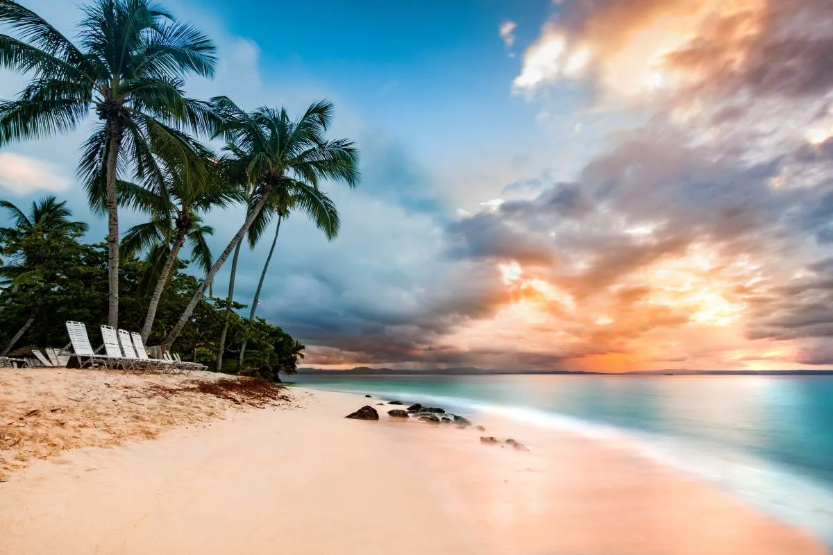 Tropical beach with palm trees and lounge chairs on sandy shore at sunset, with dramatic clouds in the sky and calm sea reflecting vibrant orange and blue colors.