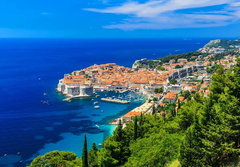 Aerial view of Dubrovnik, Croatia, showing the historic old town with orange rooftops, stone walls, and a small harbor, surrounded by deep blue sea and lush green hills under a bright blue sky.