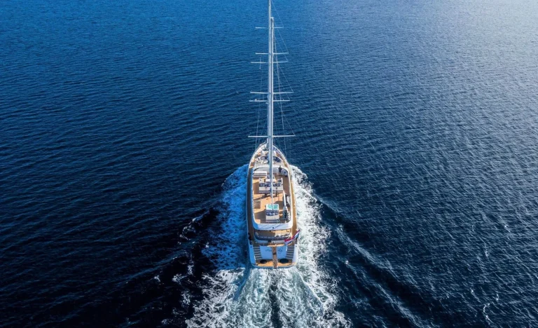 Aerial view of a luxurious yacht sailing on deep blue water, leaving a foamy wake behind as it moves forward under a clear sky.
