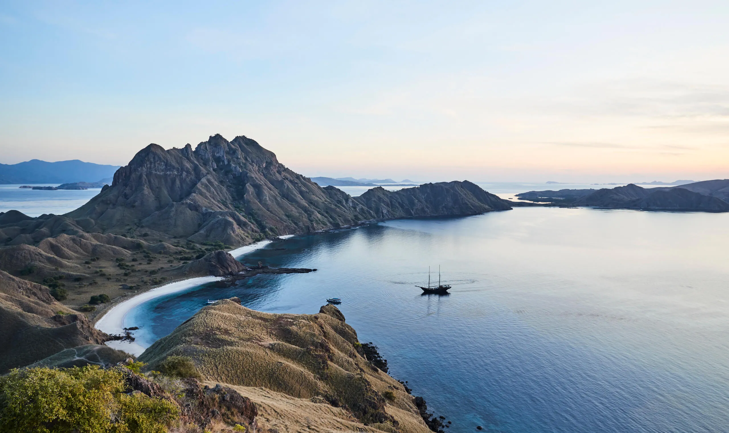 A panoramic view of a curved coastline with rugged hills and mountains, surrounded by calm blue water. A sailboat floats in the bay under a clear sky at sunset or sunrise.