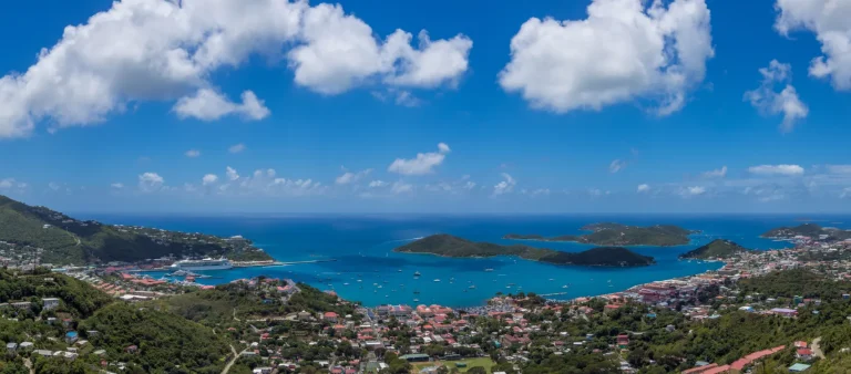 A panoramic view of a coastal town with red-roofed buildings, lush green hills, and a bright blue bay dotted with boats and small islands under a sunny, partly cloudy sky.