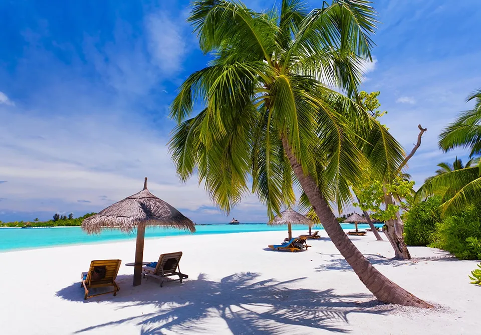 A tropical beach with white sand, turquoise water, palm trees, and lounge chairs under thatched umbrellas on a sunny day with a blue sky and scattered clouds.