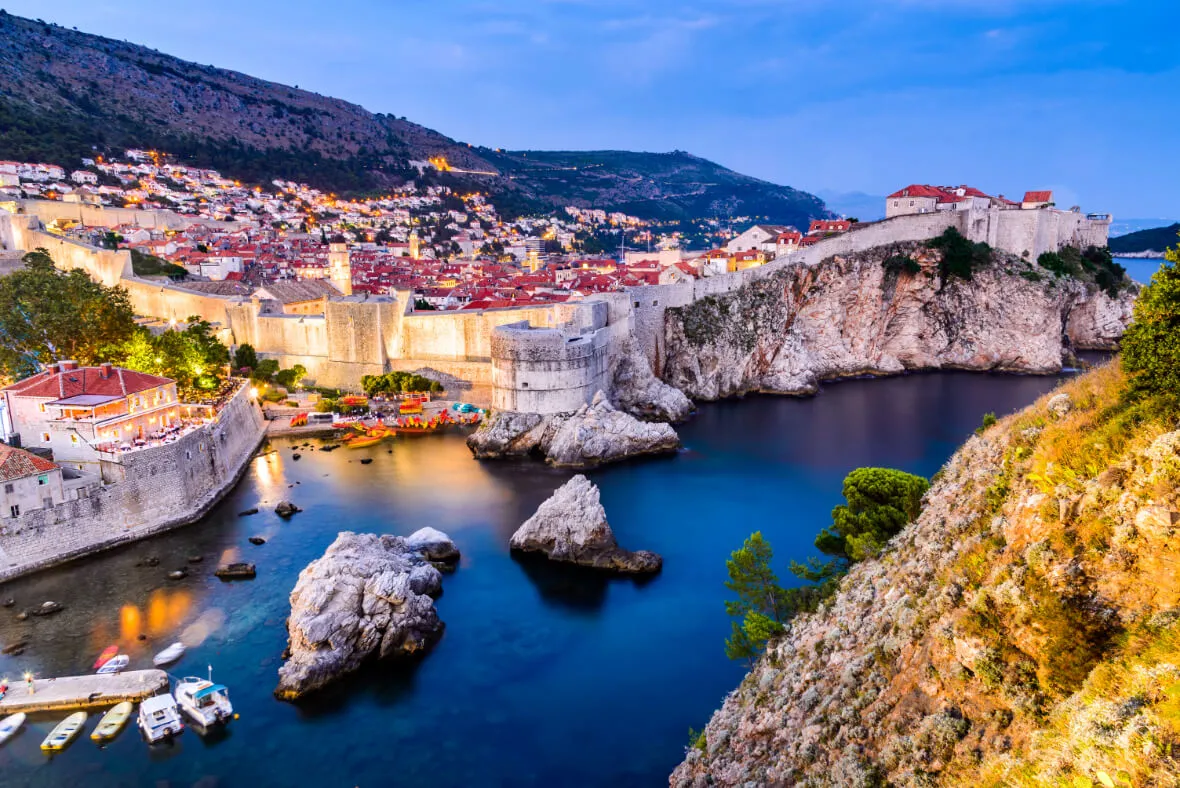 Coastal view of Dubrovnik, Croatia at dusk, featuring historic stone city walls, illuminated buildings, rocky cliffs, and calm blue water with small boats near the shore.