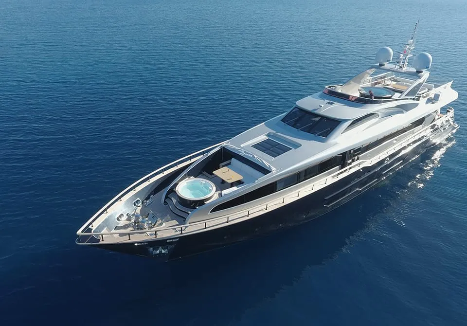 A large luxury yacht with a hot tub on the deck floats on calm blue water, viewed from above. The yacht features sleek, modern design and spacious outdoor areas.