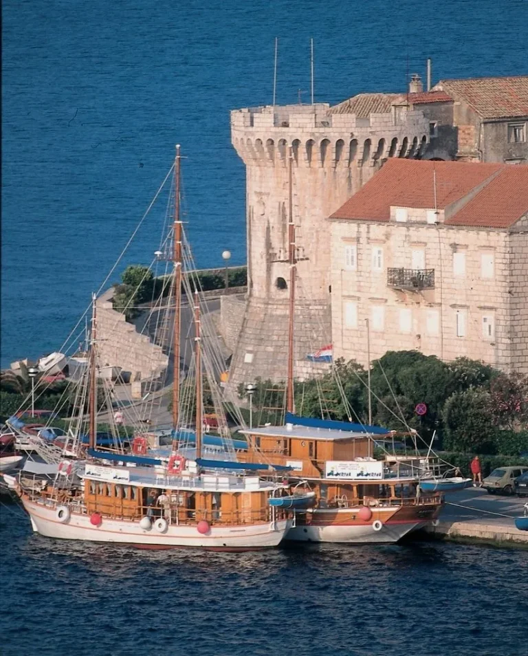 Two wooden sailboats are docked by a stone waterfront, with a historic round tower and old stone buildings in the background, overlooking a deep blue sea under clear skies.