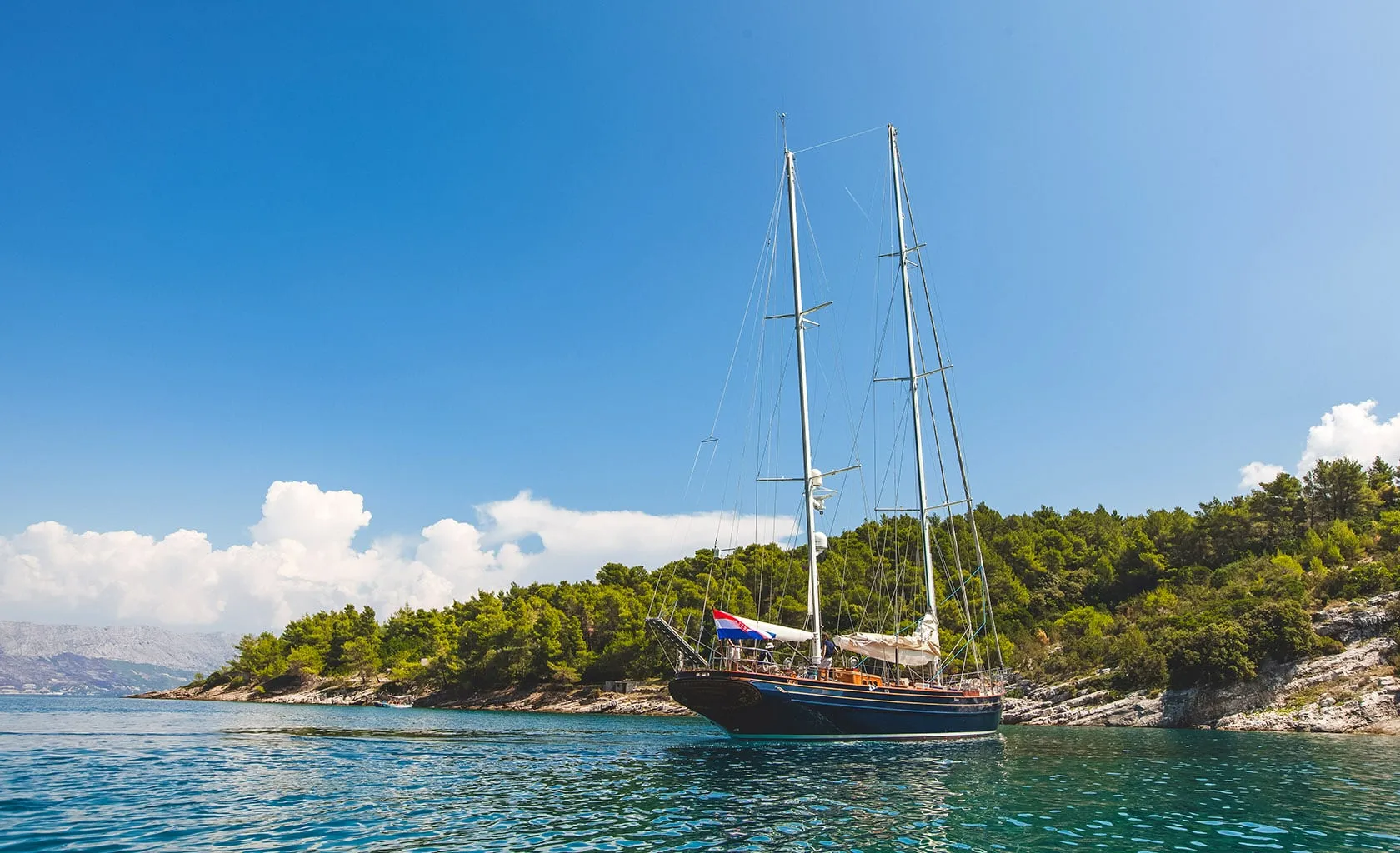 A sailboat with two tall masts floats on calm blue water near a forested, rocky shoreline under a bright blue sky with scattered white clouds.