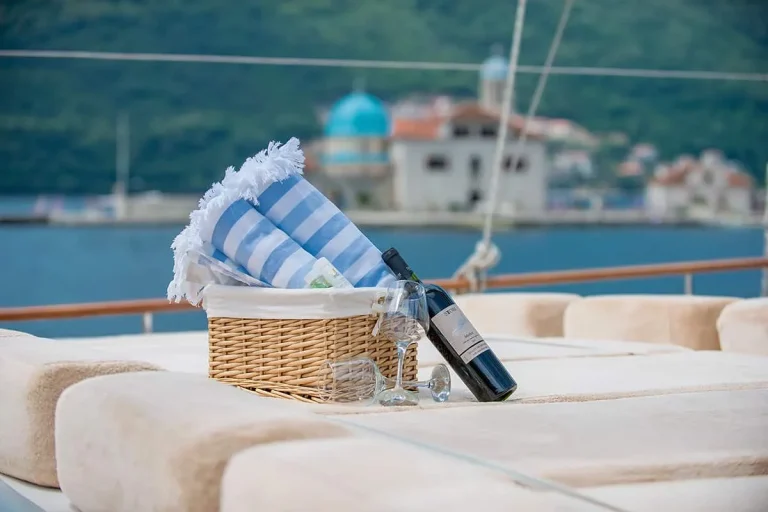 A wicker basket with blue and white striped towels, a bottle of wine, and two wine glasses sits on cushioned seating of a boat, with water and a building with a blue dome in the background.