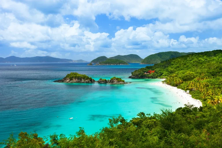 A tropical beach with turquoise water, white sand, and lush green hills under a partly cloudy sky. Small islands and distant hills are visible on the horizon.