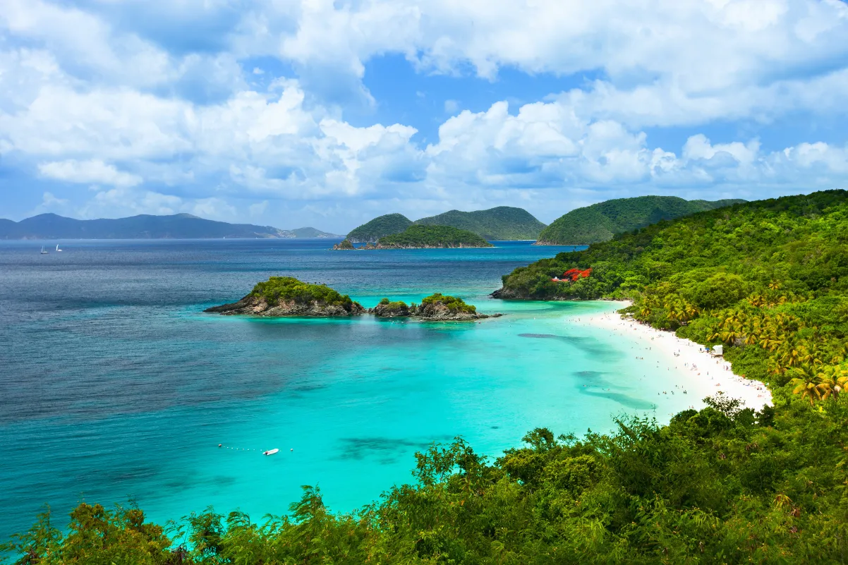 A tropical beach with turquoise water, white sand, and lush green hills under a partly cloudy sky. Small islands and distant hills are visible on the horizon.