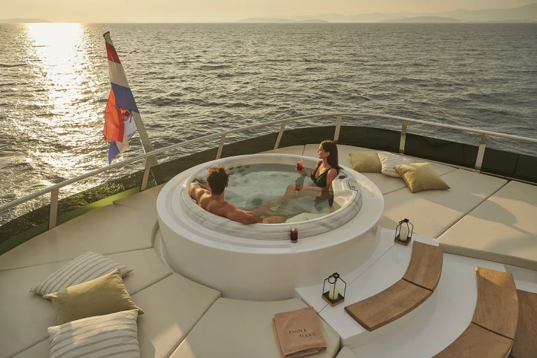 A man and woman relax in a hot tub on a yacht at sunset, holding drinks. Cushions, a book, lanterns, and a flag decorate the deck, with the calm sea and distant mountains in the background.