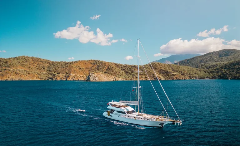 A white sailboat with tall masts sails on deep blue water near a rugged, green coastline under a sunny, clear sky with a few scattered clouds. Hills and mountains rise in the background.