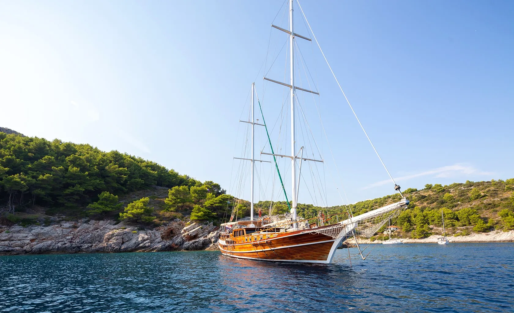 A large wooden sailboat with tall masts floats on calm blue water near a rocky, tree-covered shoreline under a clear sky.
