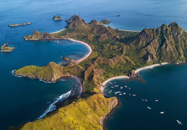 Aerial view of a lush, green island with rugged hills, surrounded by blue ocean. Several curved beaches and boats are visible near the coastline. Small islands can be seen in the distance under a clear sky.