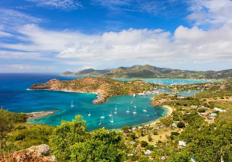 Aerial view of a tropical bay with turquoise water, sandy shores, anchored sailboats, green hills, and scattered houses under a partly cloudy blue sky.