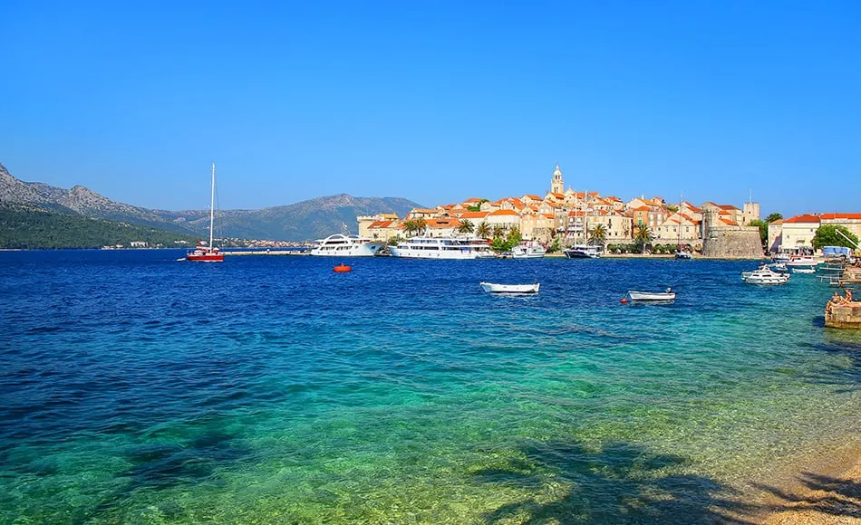 A small coastal town with orange-roofed buildings sits beside clear blue water. Boats are anchored near the shore, and green hills rise in the background under a bright blue sky.