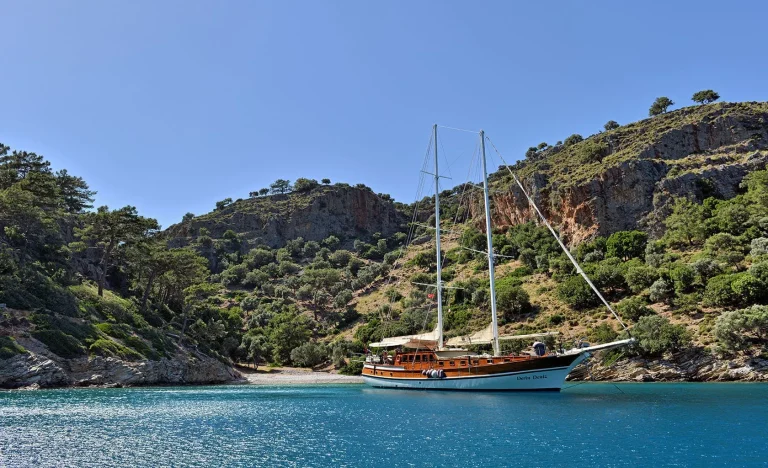 A wooden sailboat is anchored in calm turquoise water near a small, secluded beach, surrounded by rocky hills and lush green trees under a clear blue sky.