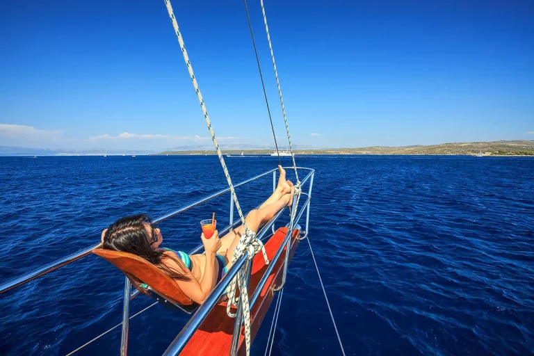 A woman lounges on a chair at the bow of a sailboat, holding a drink and relaxing under a clear blue sky with deep blue water and distant land visible in the background.