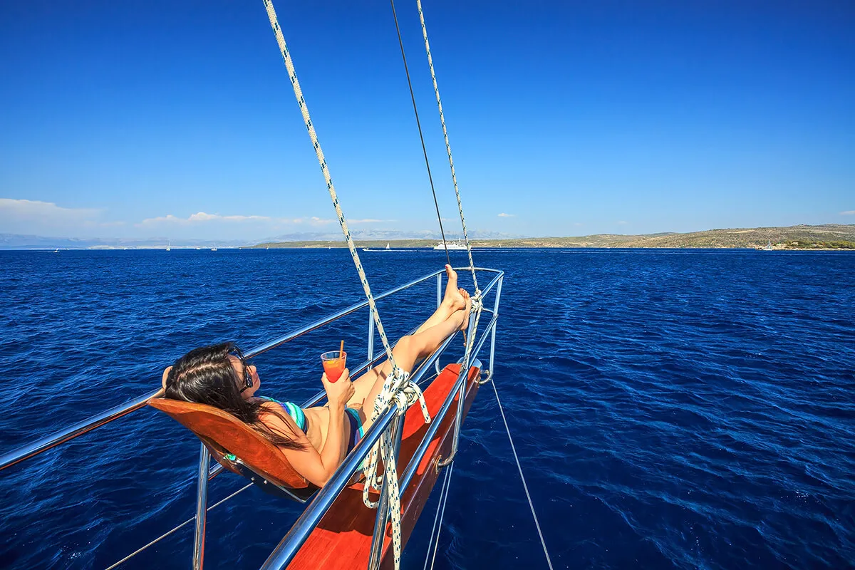A woman lounges on a chair at the bow of a sailboat, holding a drink and relaxing under a clear blue sky with deep blue water and distant land visible in the background.
