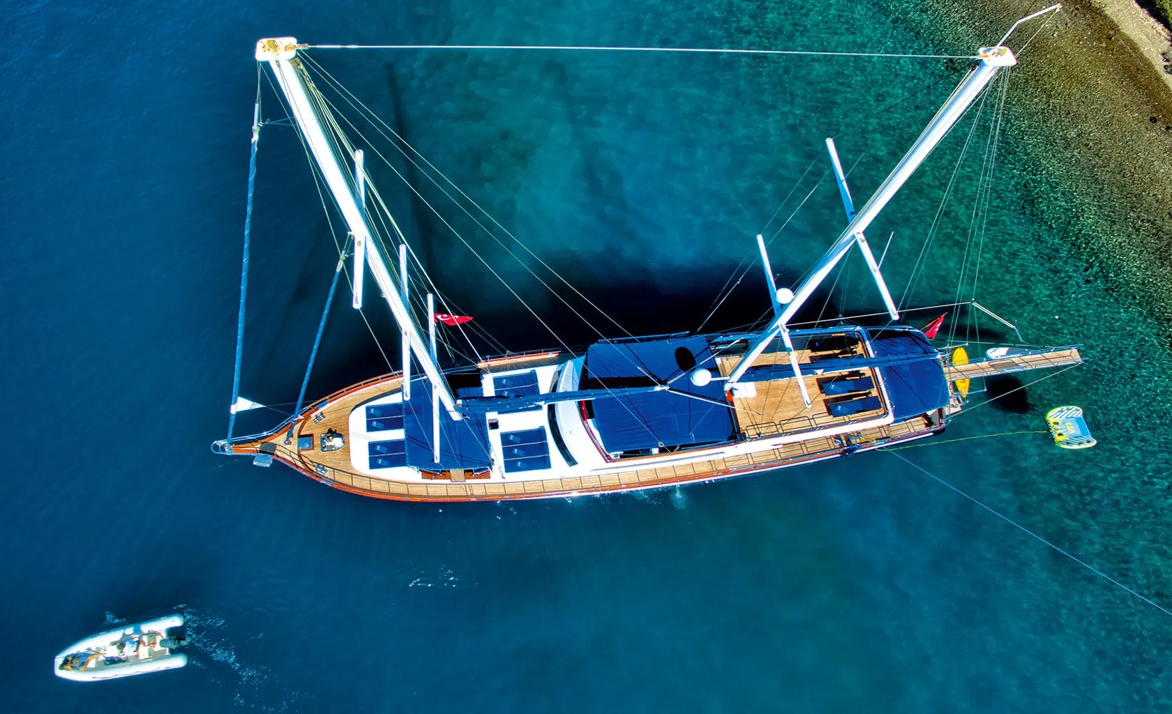 Aerial view of a large sailboat with blue canopies anchored near a coastline in clear blue water. A small motorboat approaches the sailboat on the left side of the image.