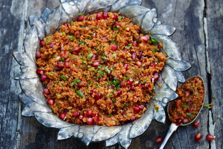 A decorative silver bowl filled with reddish quinoa salad, garnished with fresh herbs and pomegranate seeds, sits on a rustic wooden surface with a spoonful of salad placed beside the bowl.