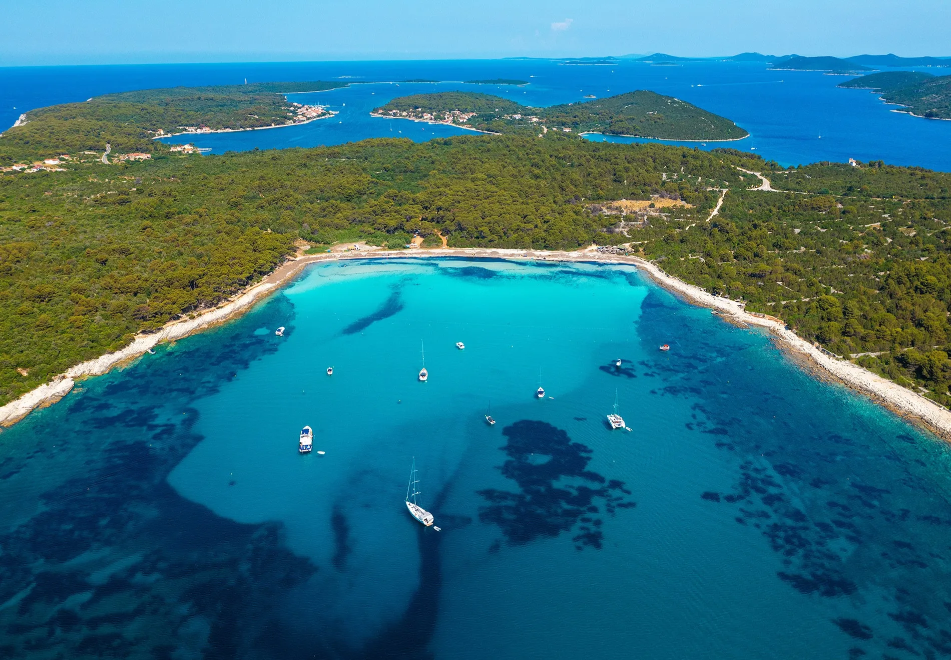 Aerial view of a turquoise bay with anchored sailboats surrounded by dense green forest and several islands, under a clear blue sky.