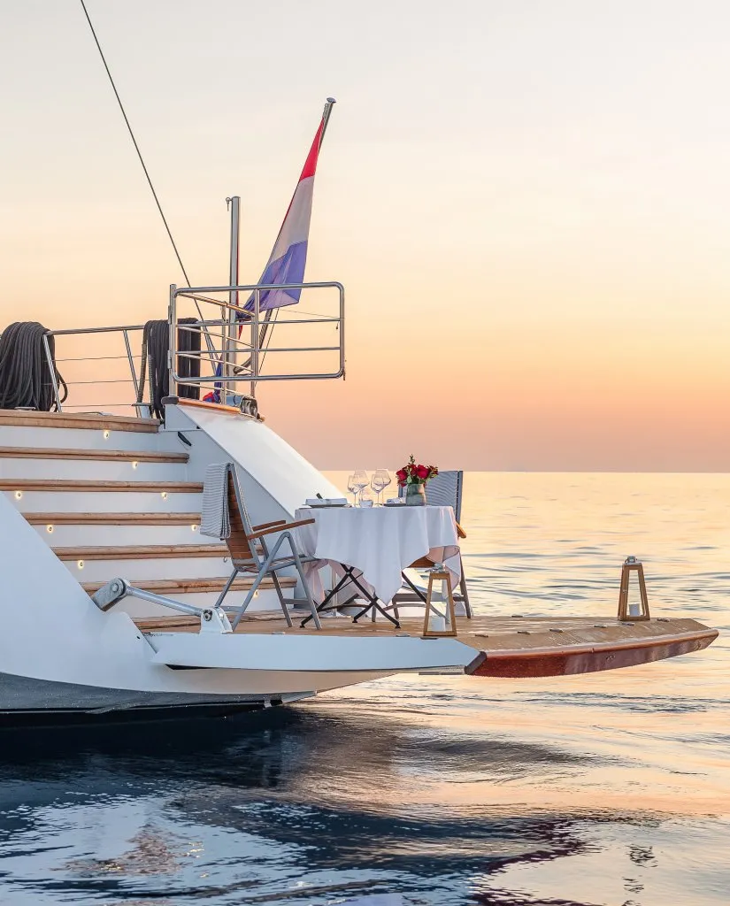 A small dining table with two chairs is set on the deck of a yacht at sunset. Wine glasses, flowers, and a Dutch flag are visible, with calm sea and a pastel sky in the background.