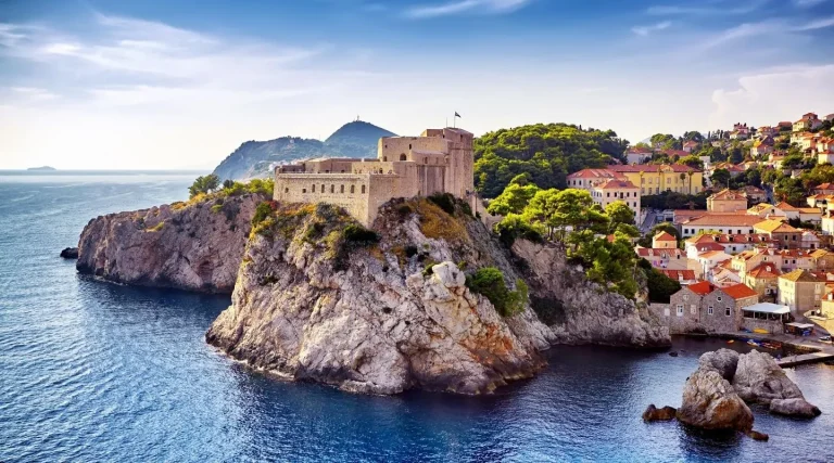 A stone fortress sits atop a rocky cliff overlooking the blue sea, with a coastal town of red-roofed buildings and green trees in the background under a clear, sunny sky.