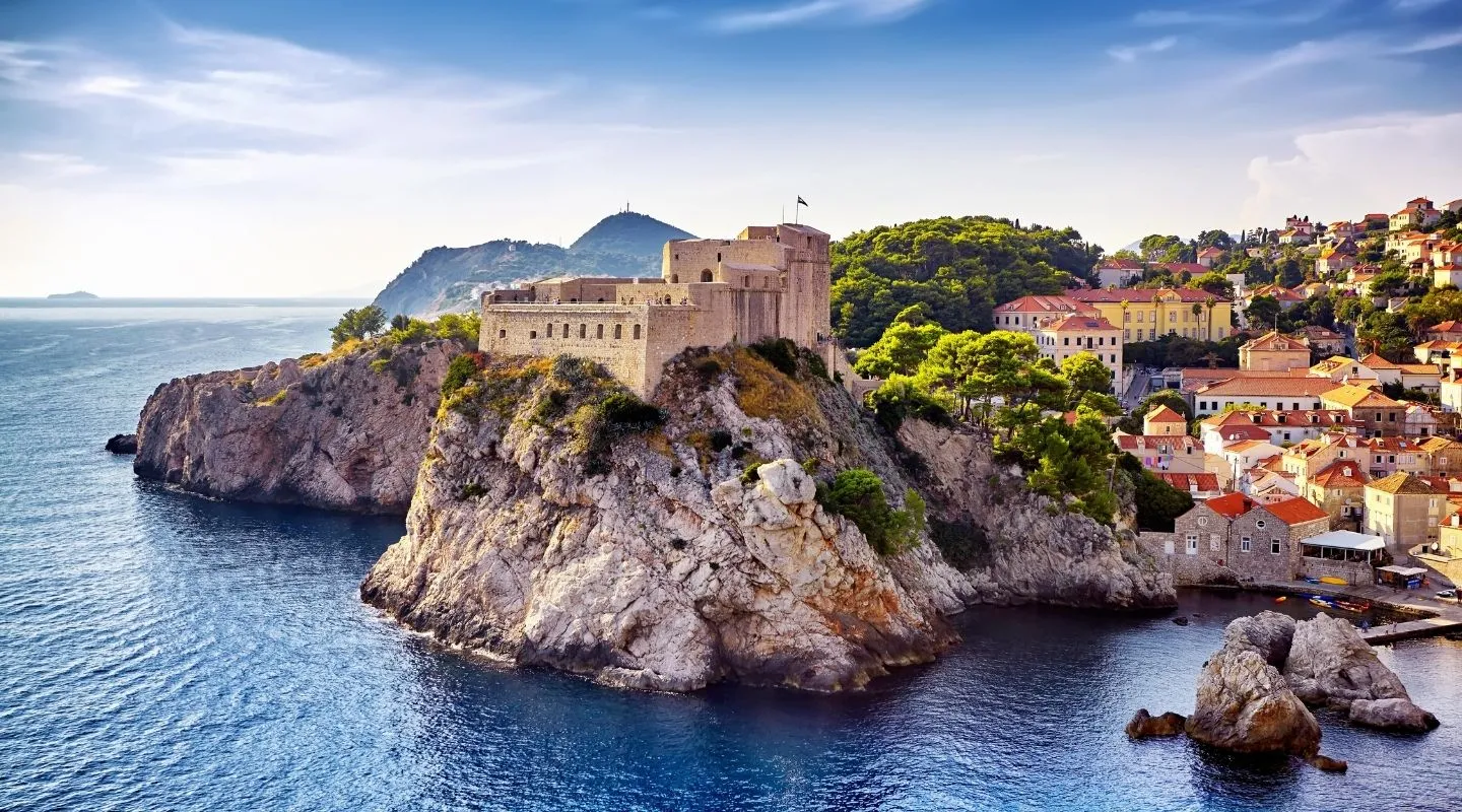 A stone fortress sits atop a rocky cliff overlooking the blue sea, with a coastal town of red-roofed buildings and green trees in the background under a clear, sunny sky.