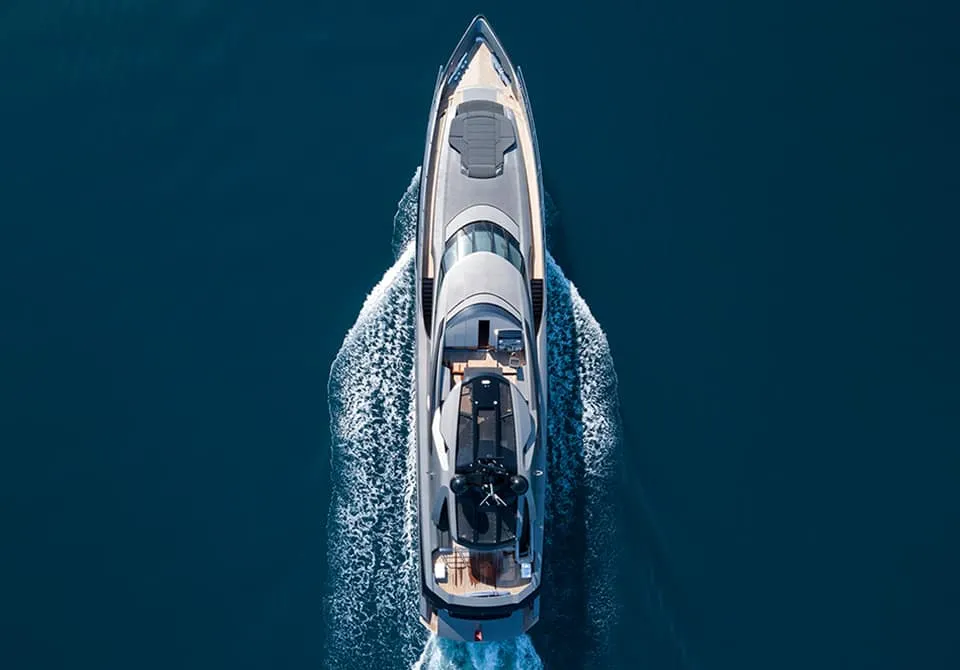Aerial view of a luxury yacht cruising through calm blue water, leaving white trails behind as it moves forward, with detailed decks and a sleek modern design visible.
