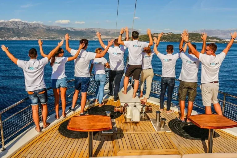 A group of people in matching shirts stand at the back of a boat, facing the water with their arms raised. The scene is set against a backdrop of blue sea and distant mountains under a clear sky.