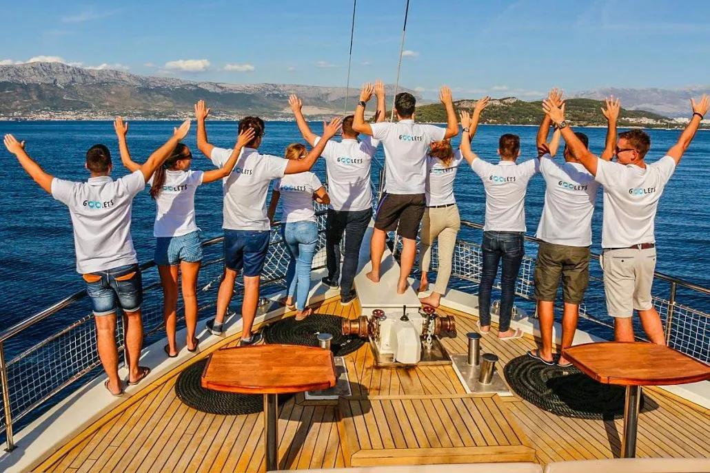A group of people in matching shirts stand at the back of a boat, facing the water with their arms raised. The scene is set against a backdrop of blue sea and distant mountains under a clear sky.