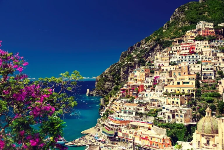 Colorful buildings cling to a steep hillside overlooking the deep blue sea in Positano, Italy, with vibrant pink flowers in the foreground and boats docked along the shore under a clear sky.