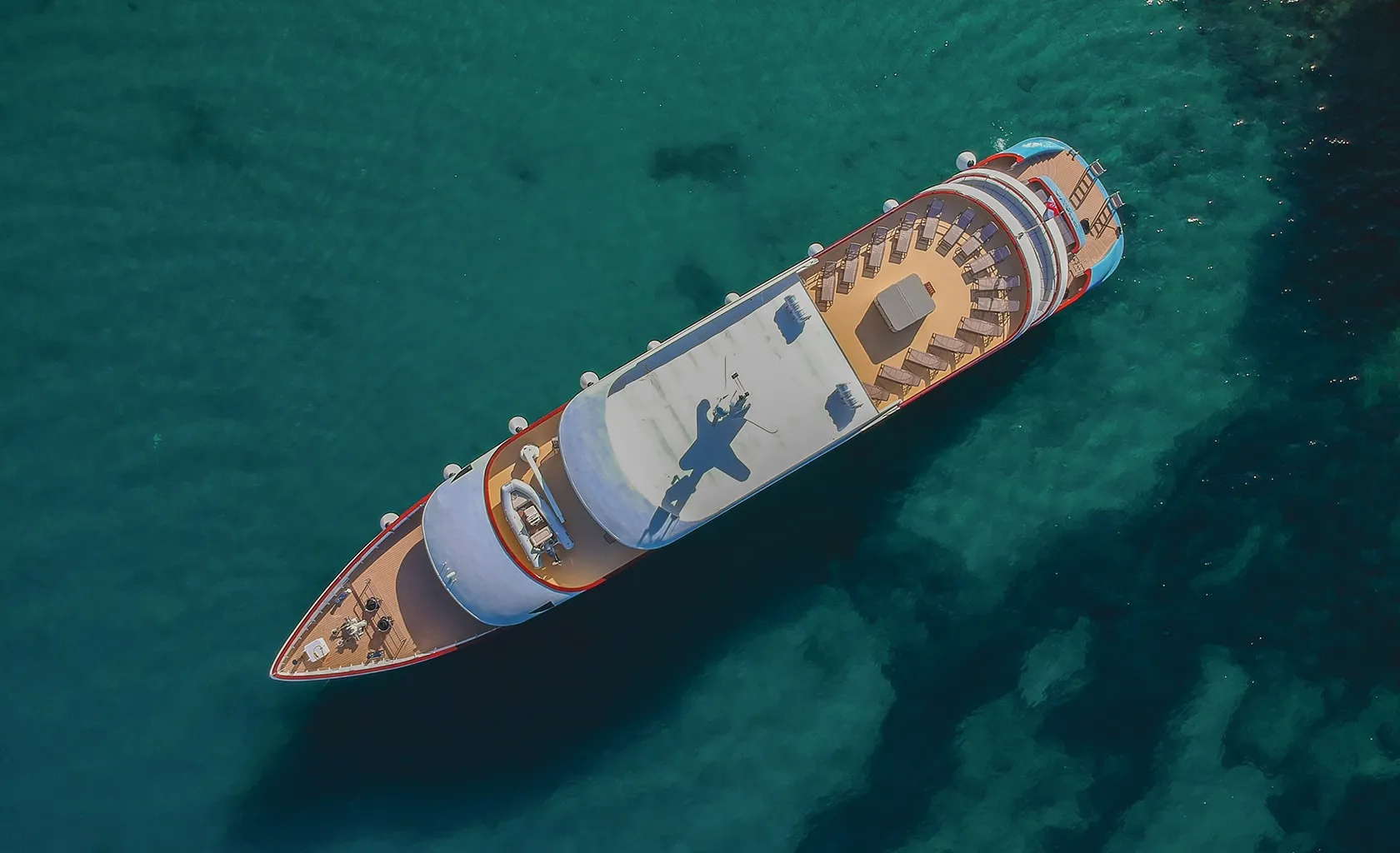 Aerial view of a cruise ship with outdoor seating areas sailing through clear turquoise water, casting a shadow on its deck and the sea below.