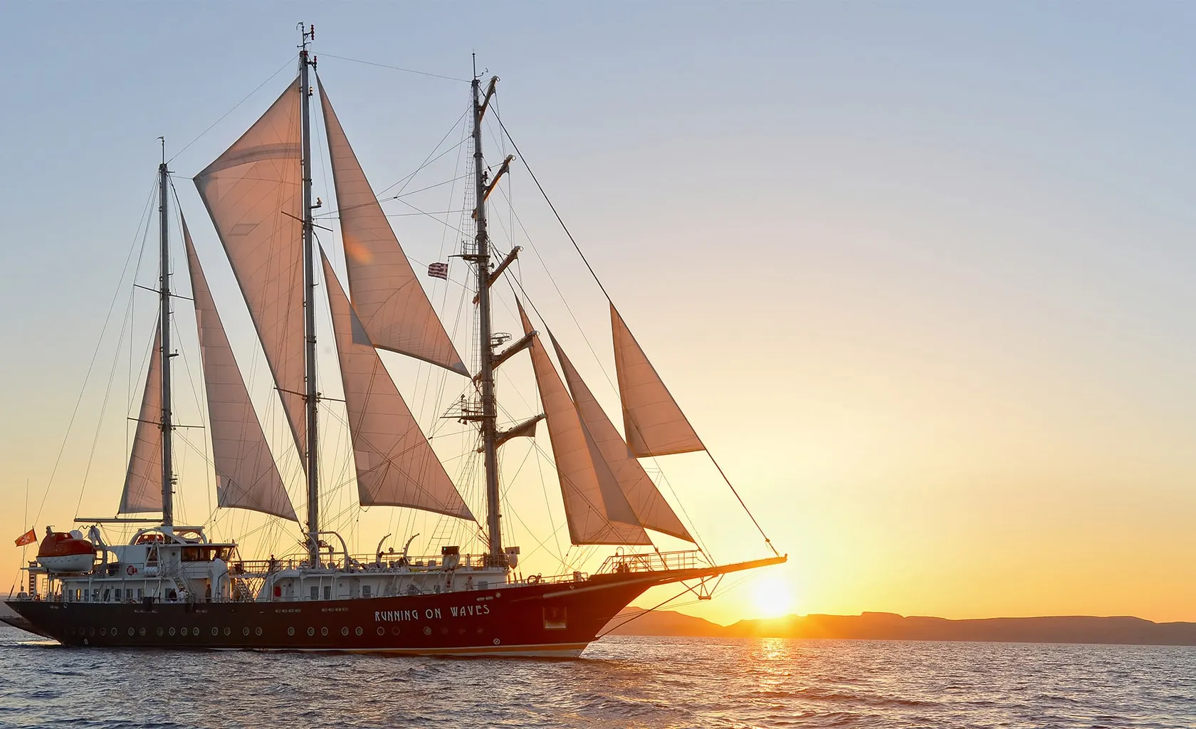 A large sailboat with white sails glides on calm water at sunset, with the sky glowing orange and the sun setting behind distant hills.