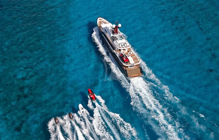 Aerial view of a large white yacht cruising on bright blue water, with three jet skis and a red speedboat creating white trails as they follow closely behind.
