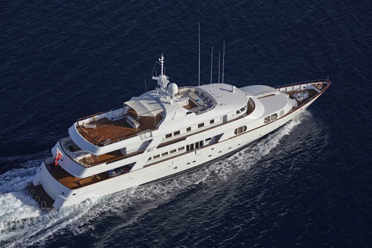 A large white luxury yacht with wooden decking sails through deep blue water, creating waves along its sides. The yacht has multiple decks and outdoor lounging areas.
