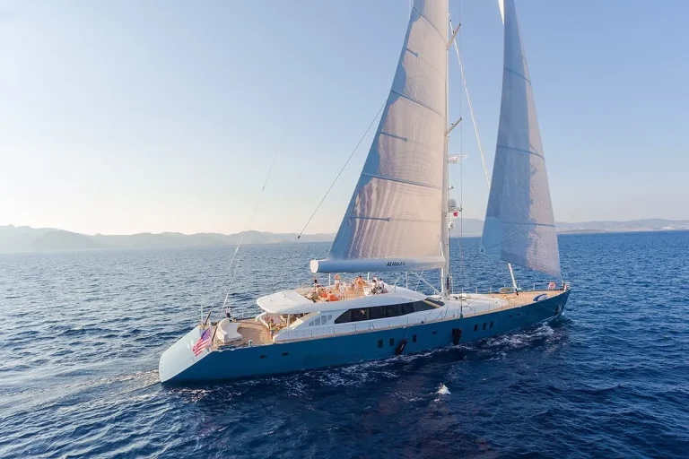 A large luxury sailboat with white sails glides across calm blue ocean water under a clear sky, with several people relaxing on its spacious deck.
