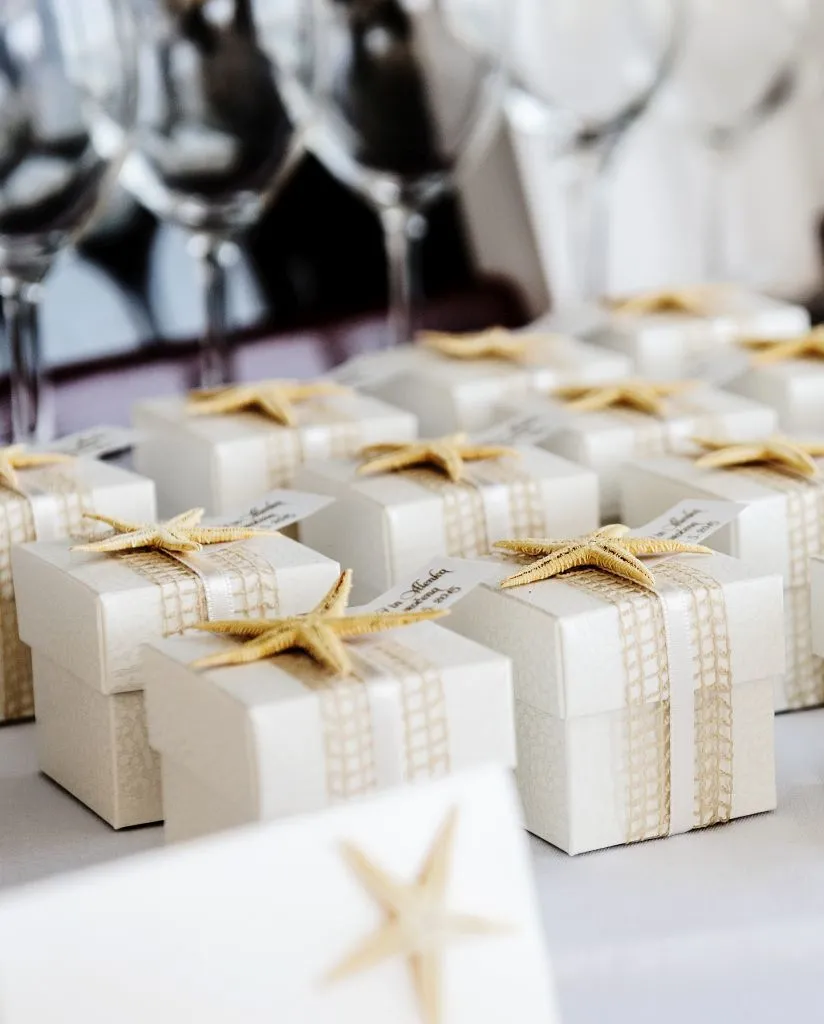 Small white gift boxes with ribbon and starfish decorations are arranged neatly on a table, with wine glasses blurred in the background.